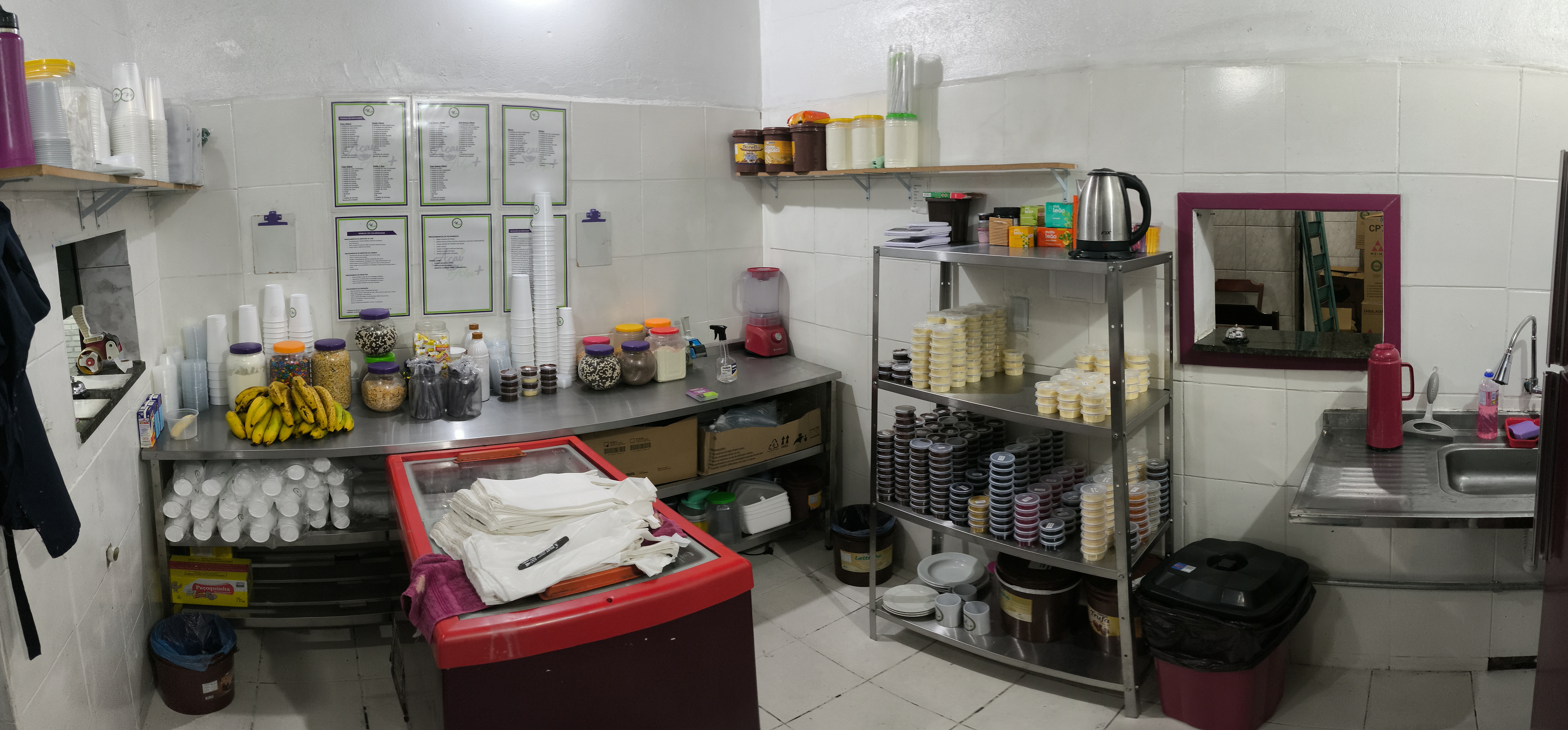 Cozinha da loja em estado maduro: bancada inox curva abastecida com 19 toppings, pilhas de copos, freezer e prateleira inox com potes pré-porcionados pra delivery